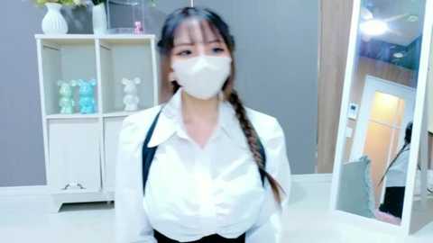 Video of an Asian woman in a white shirt, black suspenders, and a white face mask, standing in a modern, bright room with a white cabinet and a mirror.