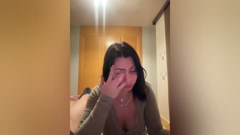 A video of a woman with medium-length black hair, wearing a beige top, covering her face with her right hand, standing in a hallway with light wooden doors and beige walls.