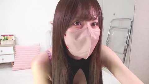 Video of an Asian woman with long black hair, wearing a pink face mask, seated at a white desk in a room with white walls and a metal ladder.