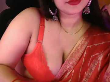 Video of a South Asian woman with fair skin, wearing a bright red saree and matching bra, adorned with large, dangling earrings. The background is blurred.