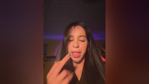 Video of a young woman with long black hair, wearing a black top, licking a finger seductively. Background shows dimly lit room with purple lighting and a bed with red sheets.