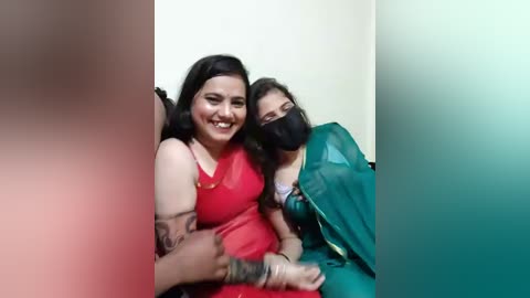 Video of two smiling women in traditional green saris, one with a mask, seated together against a plain wall, with a blurred figure in the foreground.