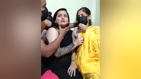 A video captures a distressed woman with a tattooed arm in a black dress, being comforted by two women in yellow saris. The background is a plain beige wall.