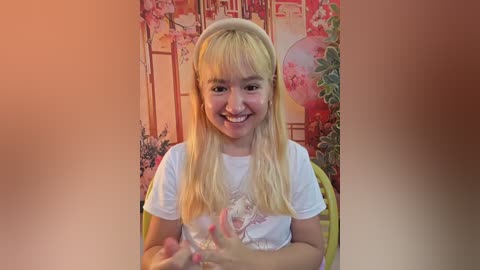 A video of a young blonde girl with a white headband, smiling, wearing a white shirt, sitting in front of a pink floral backdrop with a balloon.
