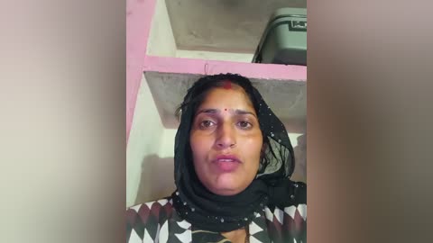 Video of a woman with medium brown skin, wearing a black hijab with white geometric patterns, and a red dot on her forehead, standing in a bathroom with white walls and a pink shelf.