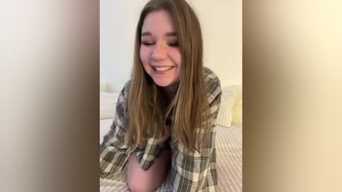 Video of a young woman with long, light brown hair, wearing a plaid shirt, kneeling on a bed with a striped sheet, smiling and looking down.