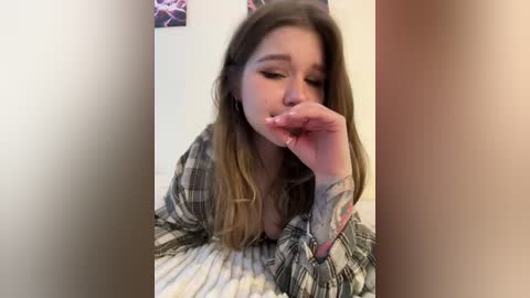 Video of a young woman with long brown hair, wearing a plaid shirt, covering her mouth with her hand, leaning against a beige wall with a blurred, colorful abstract painting.