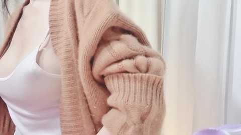 Video of a woman with a medium build, wearing a white tank top and a beige knitted cardigan. She is covering her mouth with her hand, suggesting nervousness or discomfort. Background shows a white curtain.