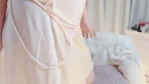 Video of a person wearing a light pink, flowy dress with a sheer, crisscrossing back, standing in a softly lit room with white curtains and a fluffy white blanket.
