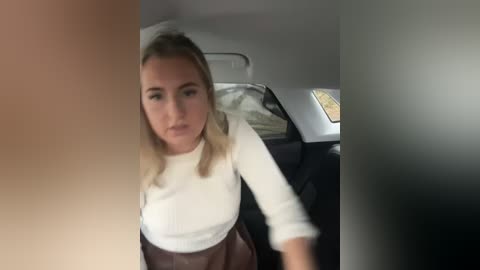 Media: Video of a blonde woman with shoulder-length hair, wearing a white long-sleeve shirt and brown skirt, driving a car. Background includes a black steering wheel, white headrest, and a window with a yellow light.