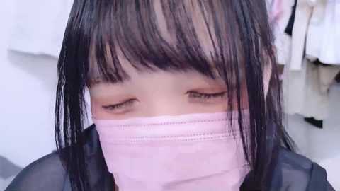 Video of a young Asian woman with long black hair and bangs, wearing a pink surgical mask and black jacket, with blurred white clothing in the background.