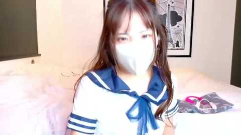 Video of an East Asian woman with long brown hair, wearing a white surgical mask and a blue sailor school uniform, seated on a bed in a dimly lit room.