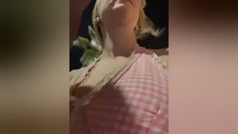 Video of a blonde woman with fair skin, wearing a pink and white checkered dress, lying on a bed with dark sheets, partially visible.