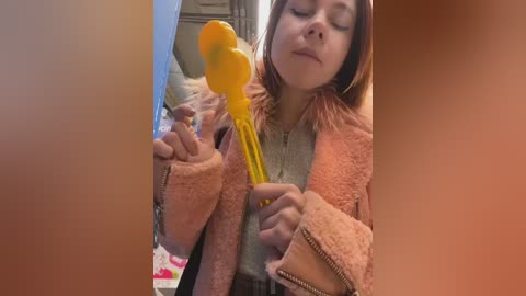 A video of a young woman with light skin and shoulder-length brown hair, wearing a pink fleece jacket, holding a yellow, duck-shaped bath toy in a bathroom with a mirror reflection.