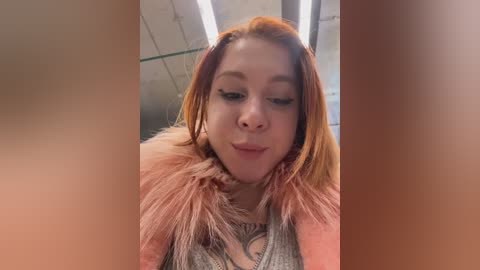 Video of a young woman with fair skin, red hair, and a pink fur-lined jacket. She has a playful expression and is in an indoor setting with fluorescent lights and a tiled ceiling.