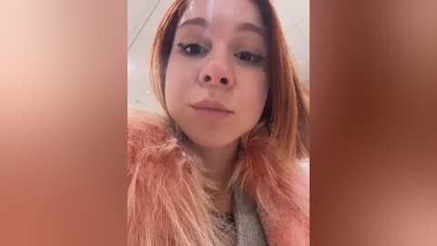 A close-up video of a young woman with fair skin, wearing a pink fur-lined coat, has shoulder-length, straight, reddish-brown hair. Her face is neutral, and she's indoors with a blurred background.