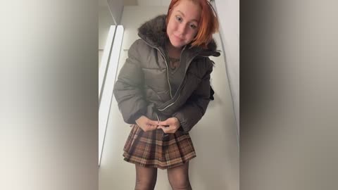 Video of a young woman with fair skin and red hair, wearing a puffy green jacket with fur collar, brown plaid skirt, and sheer black tights. She stands in a narrow hallway with white walls and fluorescent lighting.