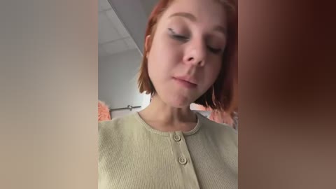 Video of a young Caucasian woman with pale skin and short red hair, wearing a beige cardigan, standing indoors. The background features a blurred, neutral-toned room.