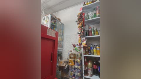 Video of a cluttered, brightly lit kitchen refrigerator with various food items, condiments, and snacks visible through the glass doors. The background includes a white tiled wall and a shelf with more food items.