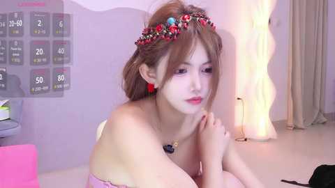 Video of a young East Asian woman with light skin, wearing a floral crown, pink dress, and red earrings, sitting contemplatively in a soft-lit room with a calendar and lamp.