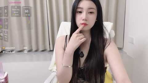 A video of an East Asian woman with long black hair, fair skin, and red lipstick, sitting on a white chair. She is wearing a black lace top and has a finger to her lips.