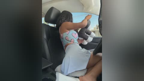 Video of a young woman with dark skin, long black hair, wearing a light blue crop top with heart patterns and a light blue skirt, sitting in a car, holding a stuffed animal.