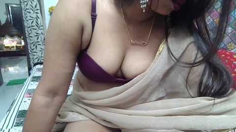 Video of a South Asian woman with medium skin tone, wearing a purple bra and beige sari, partially draped. She has long, dark hair, gold earrings, and a gold necklace. Background shows a tiled floor and colorful wall patterns.