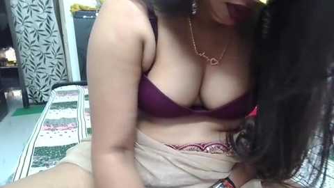 Video of a woman with medium skin tone, wearing a maroon bra and a white skirt, revealing her large breasts. She has long, dark hair and a gold necklace. Background includes a patterned rug and a black-and-white curtain.