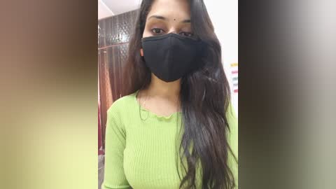 A video of a South Asian woman with long, dark hair, wearing a green ribbed top and black face mask, standing indoors with a blurred background.