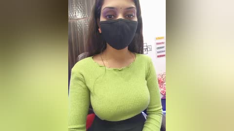 A video of a South Asian woman with medium skin tone, wearing a green ribbed sweater, black face mask, and black skirt, standing indoors. Background shows a pink and yellow wall with hanging objects.