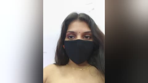 A video of a South Asian woman with long black hair, brown skin, and a black face mask, wearing a gold necklace, standing indoors against a plain white background.