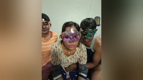 Video of three South Asian adults wearing colorful, ornate masquerade masks. The woman in the center, with medium complexion, wears a purple mask and floral-patterned blouse. The man on the left, with a light brown complexion, wears an orange-striped shirt. The man on the right, with a darker complexion, wears a green mask and a sleeveless shirt.