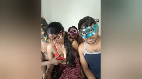 Video of four people in a room, two women with black hair and one man in a teal mask, wearing revealing red and purple outfits.