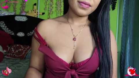 A video of a woman with long black hair, wearing a low-cut red dress, gold necklace, and pink lipstick. Background features green walls, red couch, and a black and white patterned blanket.