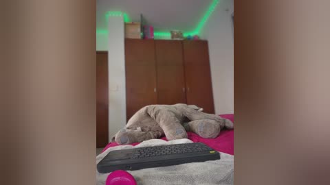 Video of a cozy bedroom with a plush brown teddy bear and a black keyboard on a bed. A green LED strip lights up the wooden wardrobe in the background.