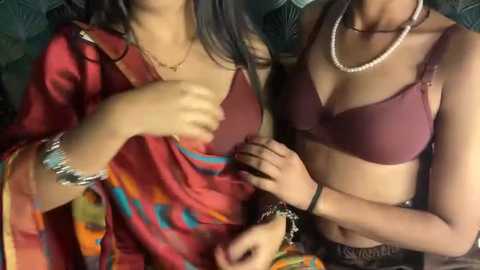 Video of two South Asian women in maroon lingerie, adjusting each other's bras. One wears a red sari with colorful patterns, the other has a pearl necklace.