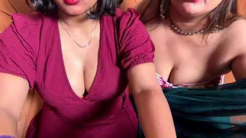 Video of two women, one in a low-cut maroon dress, the other in a green strapless top, leaning close, with visible cleavage.