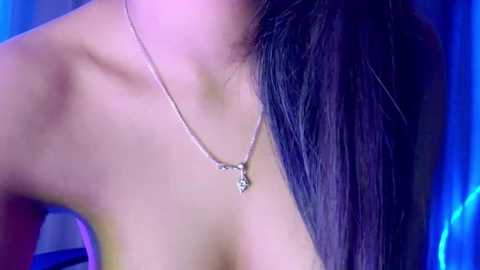 Video of a topless woman with long black hair, wearing a silver necklace with a small pendant. The background features blue and purple lighting, creating a dramatic effect.