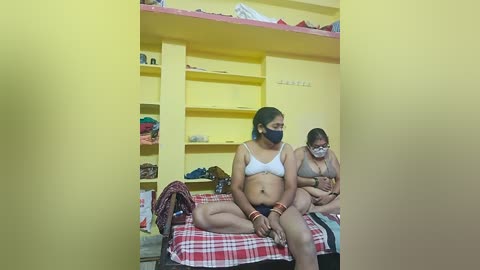 Video of two women in a small, cramped bedroom with yellow walls, wearing face masks, sitting on a red and white plaid bedspread. One woman has a white bra, and the other is topless. Shelves and clothes are visible in the background.
