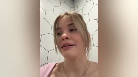 A candid video of a young, fair-skinned woman with blonde hair, wearing a pink top, standing in a bathroom with white hexagonal tile walls and a light pink towel hanging.