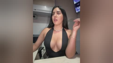 Video of a curvy, light-skinned woman with long black hair, wearing a revealing black halter dress, sitting at a desk in a dimly lit room with a monitor on the wall.