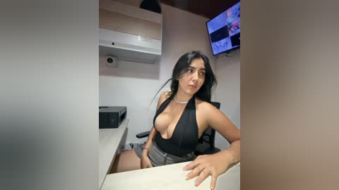 Video of a Latina woman with long black hair, medium skin tone, wearing a revealing black halter top, sitting at a desk in a minimalist office, with a TV screen displaying a news broadcast.