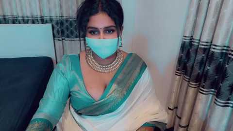 Video of a South Asian woman with medium skin tone and dark hair, wearing a teal sari with gold embroidery, a gold necklace, and a light blue mask. Background features a beige curtain and a dark couch.