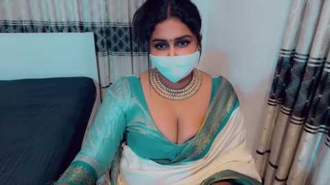 Video of a South Asian woman with medium skin tone, dark hair, wearing a teal silk sari with gold embroidery, large gold necklace, and a blue surgical mask, seated indoors on a bed with black and white striped curtains.
