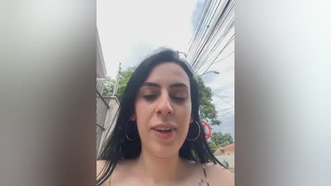 Video of a young woman with long black hair and fair skin, wearing hoop earrings, captured from a low angle, with power lines and greenery in the background.