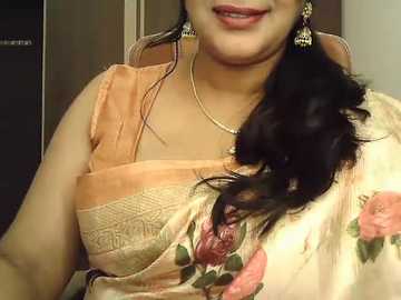 Video of a South Asian woman with medium skin tone, wearing an orange sari with floral patterns, holding a bouquet of pink and green flowers. She has long, dark hair, gold earrings, and a gold necklace.