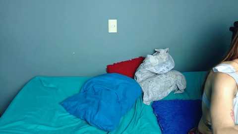 Video of a woman with light skin and long brown hair in a white tank top, sitting on a bed with turquoise sheets and pillows, surrounded by blue and red pillows and a grey blanket.