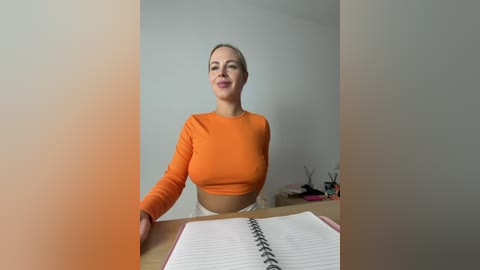 Video of a smiling, fair-skinned woman with light brown hair tied back, wearing an orange long-sleeve top, seated at a desk with a notebook in front of her.