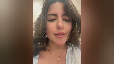 Video of a young woman with fair skin, long brown hair, and a closed-mouth smile. She wears a light grey cardigan and a gold crescent moon necklace. The background is blurred.