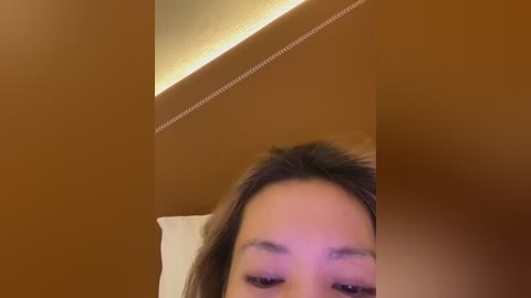 A close-up video of a woman's face with fair skin, brown eyes, and dark hair, lying on a beige pillow against a warm-toned wall.